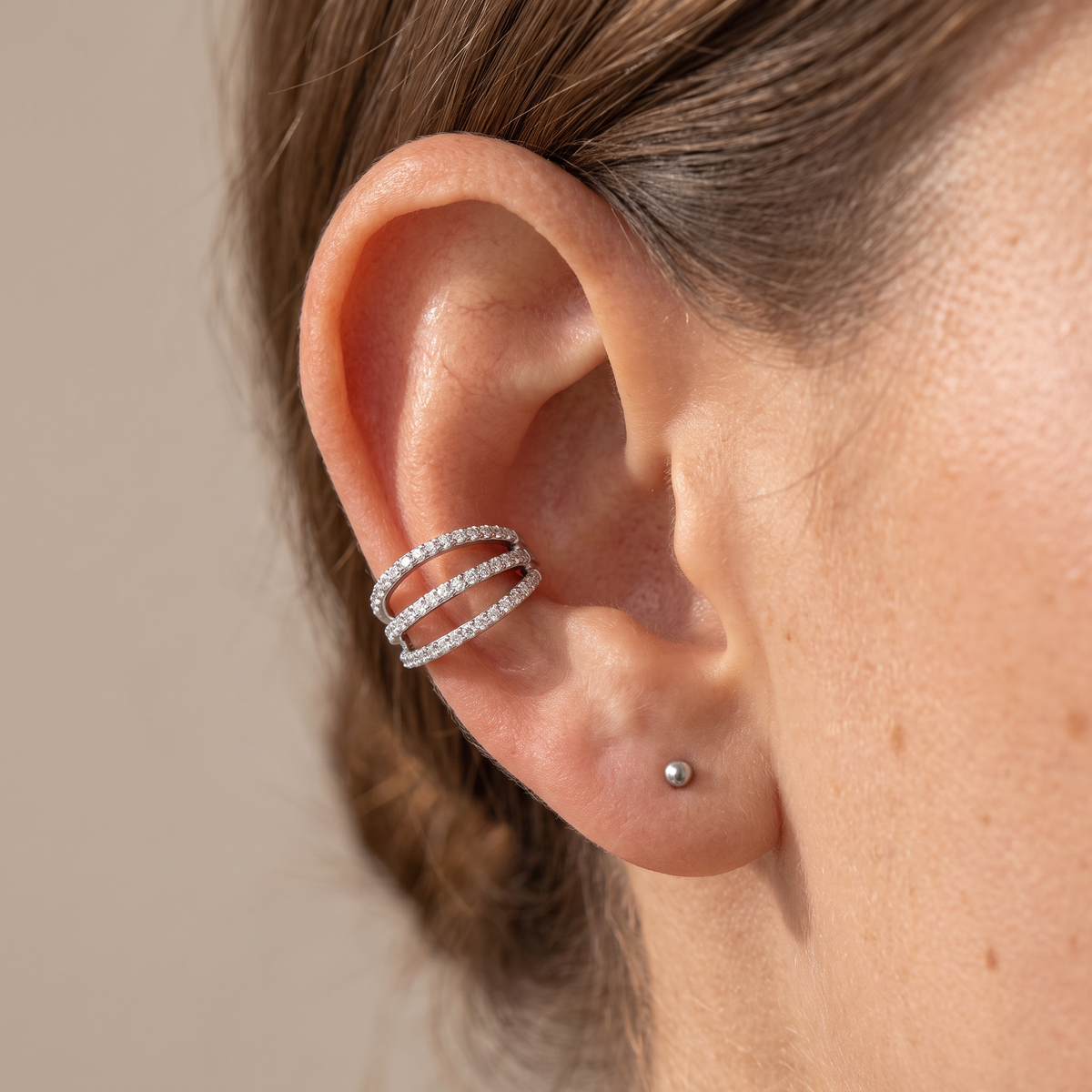 A close-up of a person's ear wearing a silver double-band ear cuff adorned with small clear stones, alongside a small silver stud earring, against a neutral beige background.