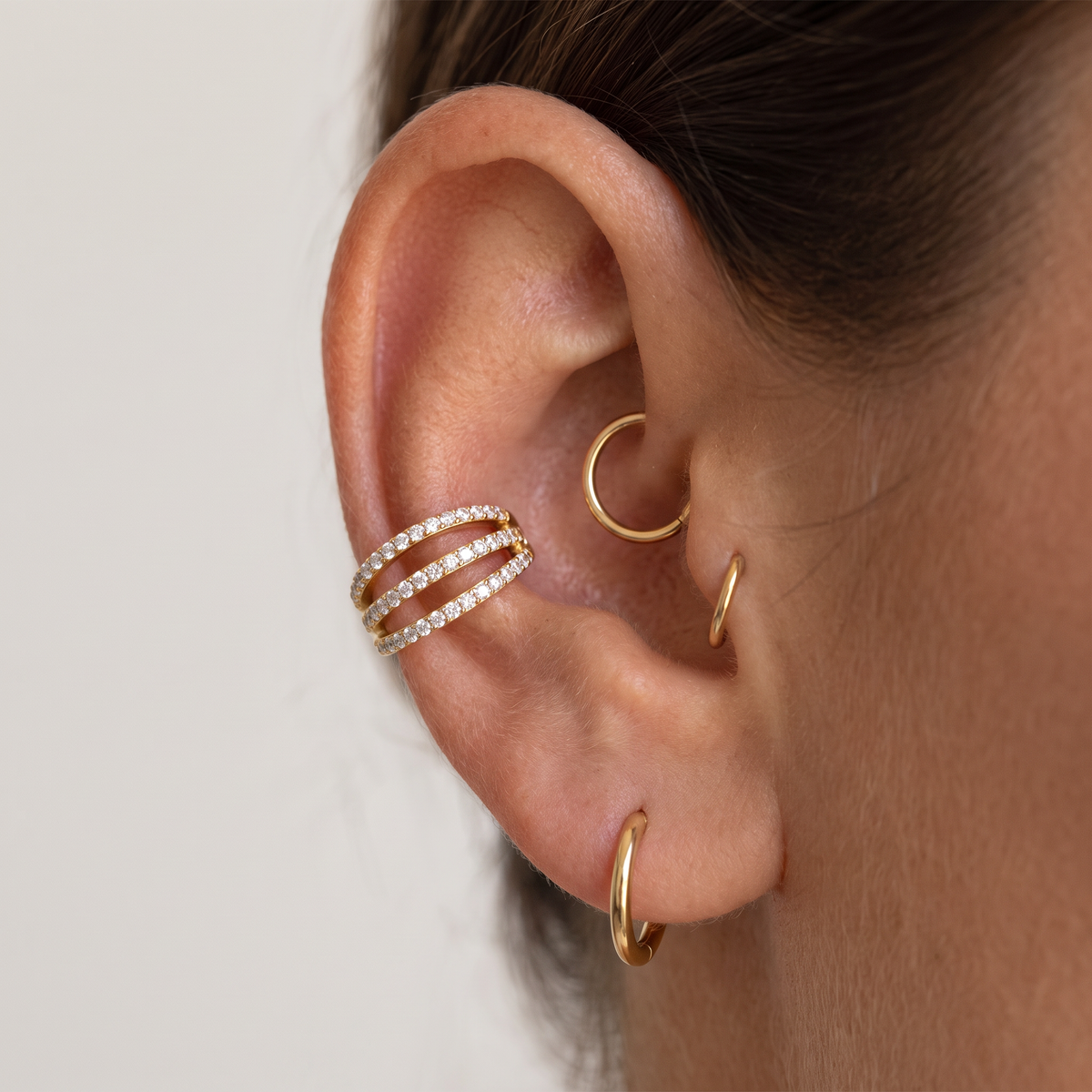 Close-up of a person's ear showcasing a stack of gold ear cuffs, including a triple-row cuff paved with clear stones and two plain gold hoop earrings.