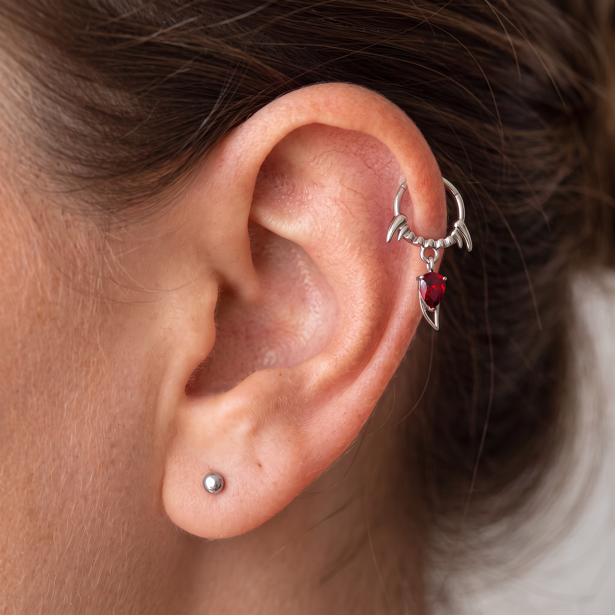 A close-up of a person's ear showcasing a silver horn-shaped lobe hoop earring adorned with a dangling red heart-shaped gemstone. A small silver stud is also visible on the earlobe.
