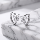 A pair of silver hoop earrings with butterfly-shaped crystal accents, displayed on a white marble block against a marble background.
