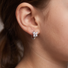 A close-up of a person's ear wearing a silver huggie hoop earring adorned with a small silver flower cluster set with clear stones.