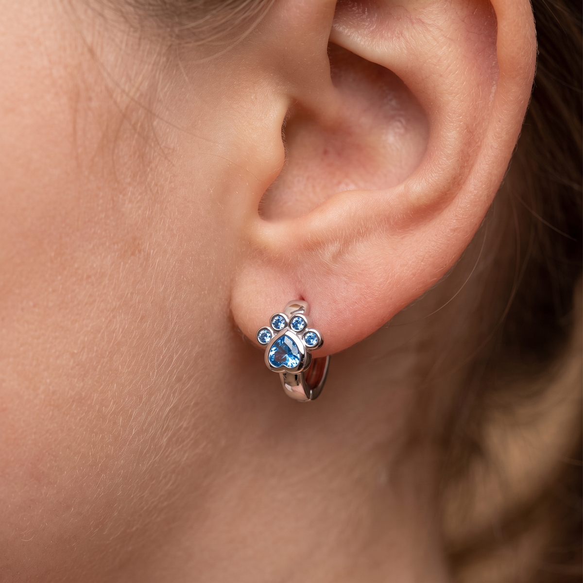A close-up of a person's ear wearing a small silver huggie hoop earring with a paw print design and blue gemstones.