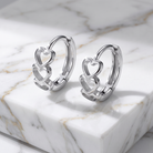 A pair of polished silver huggie hoop earrings with interlocking open heart designs, displayed on a white marble block with gold veining.