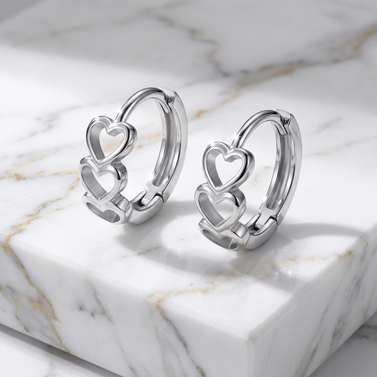 A pair of polished silver huggie hoop earrings with interlocking open heart designs, displayed on a white marble block with gold veining.