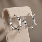 A pair of polished silver hoop earrings with sharp spikes, displayed on a beige velvet jewelry stand against a soft beige background.