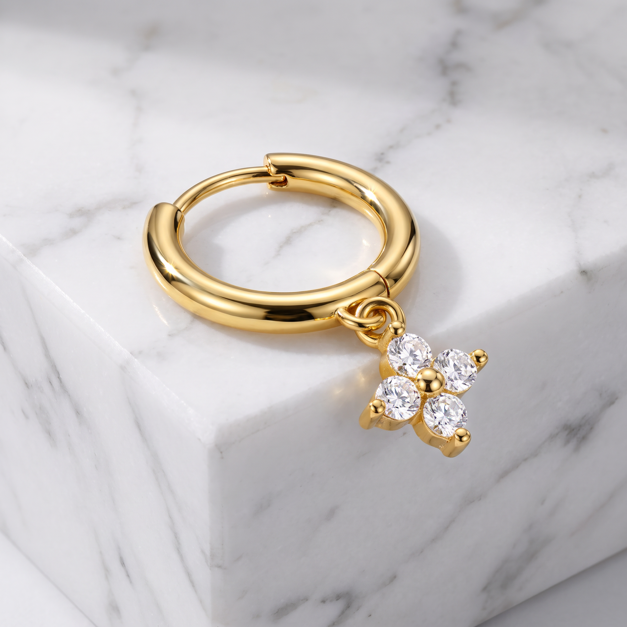 A gold huggie hoop earring with a dangling flower charm set with clear cubic zirconia stones, resting on a white marble block.
