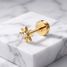 A gold starburst stud earring with a clear round-cut gemstone, displayed on a white marble block against a marble background.