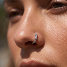 A close-up of a person's nose wearing a small silver crescent-shaped nose hoop with clear rhinestones.
