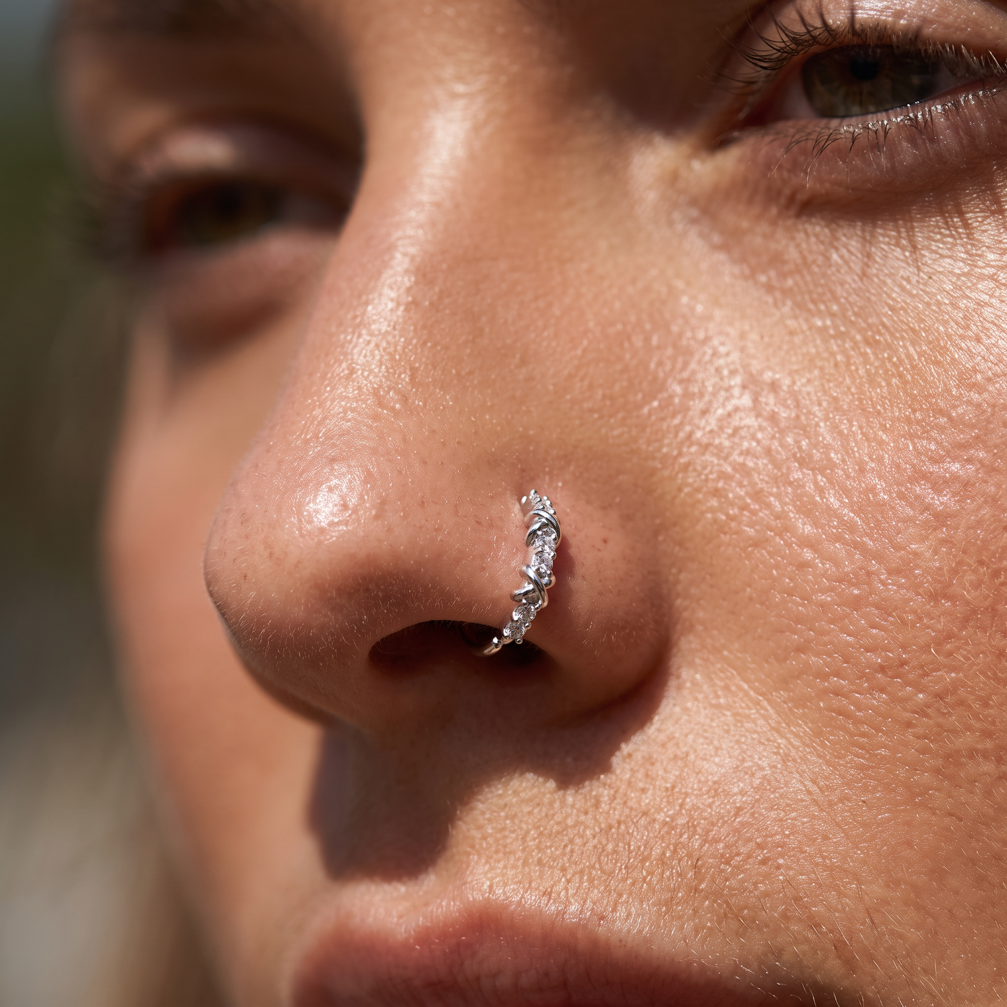 A close-up of a person's nose wearing a small silver crescent-shaped nose hoop with clear rhinestones.