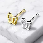 Two butterfly-shaped stud earrings displayed on a white marble surface, one in yellow gold and one in silver, both with clear crystal accents.