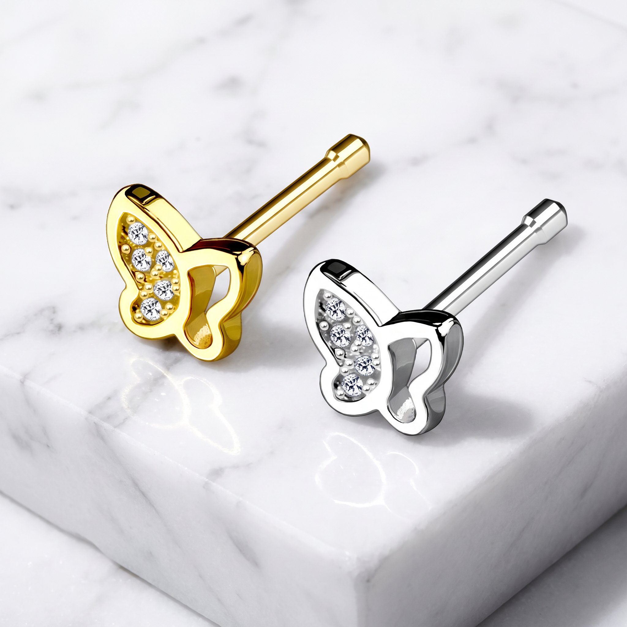 Two butterfly-shaped stud earrings displayed on a white marble surface, one in yellow gold and one in silver, both with clear crystal accents.
