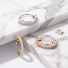 Three semi-hoop earrings displayed on a white marble surface: one silver with clear baguette stones, one rose gold with clear baguette stones, and one yellow gold with clear baguette stones. A small matching metal clasp is shown separately on the right.