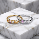Two heart-shaped stretch rings displayed on a white marble pedestal. One ring is gold with pink and clear stones, and the other is silver with pink and clear stones.