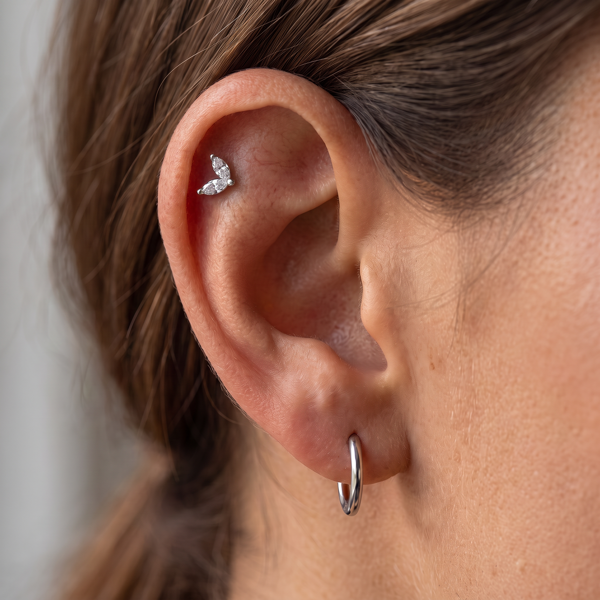 Close-up of a person's ear showcasing a small silver butterfly stud earring on the upper helix, alongside a silver hoop earring in the lobe.