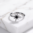 A silver ring shaped like a starburst, featuring a central black gemstone surrounded by small clear stones, displayed on a white marble block.