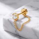 A gold stud earring with a clear round-cut gemstone and a delicate gold chain dangle, displayed on a white marble block.