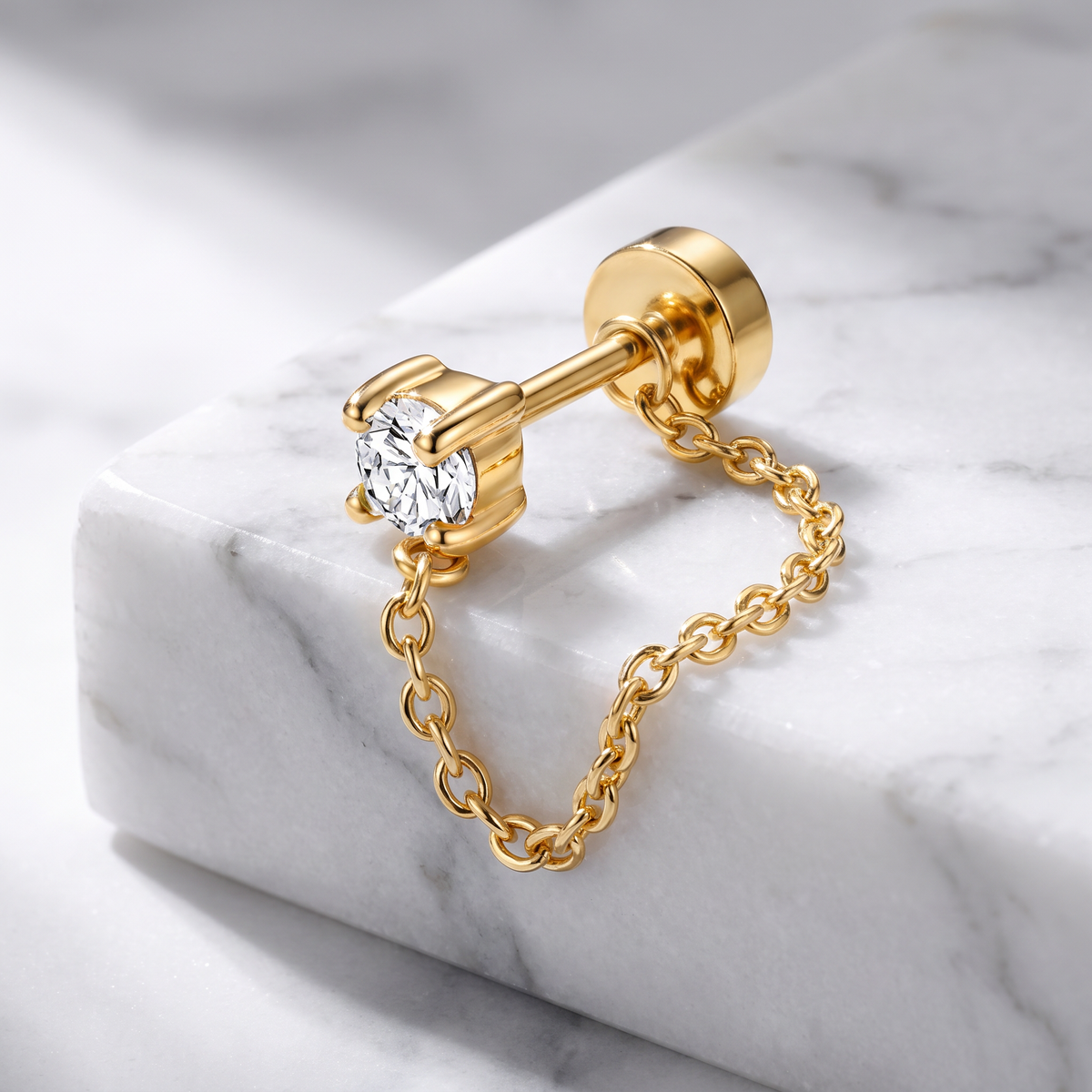 A gold stud earring with a clear round-cut gemstone and a delicate gold chain dangle, displayed on a white marble block.