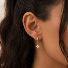 A close-up of a person's ear wearing a small gold hoop earring with a dangling floral cluster of clear stones.