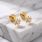 A pair of gold starburst stud earrings with clear gemstones, displayed on a white marble block.