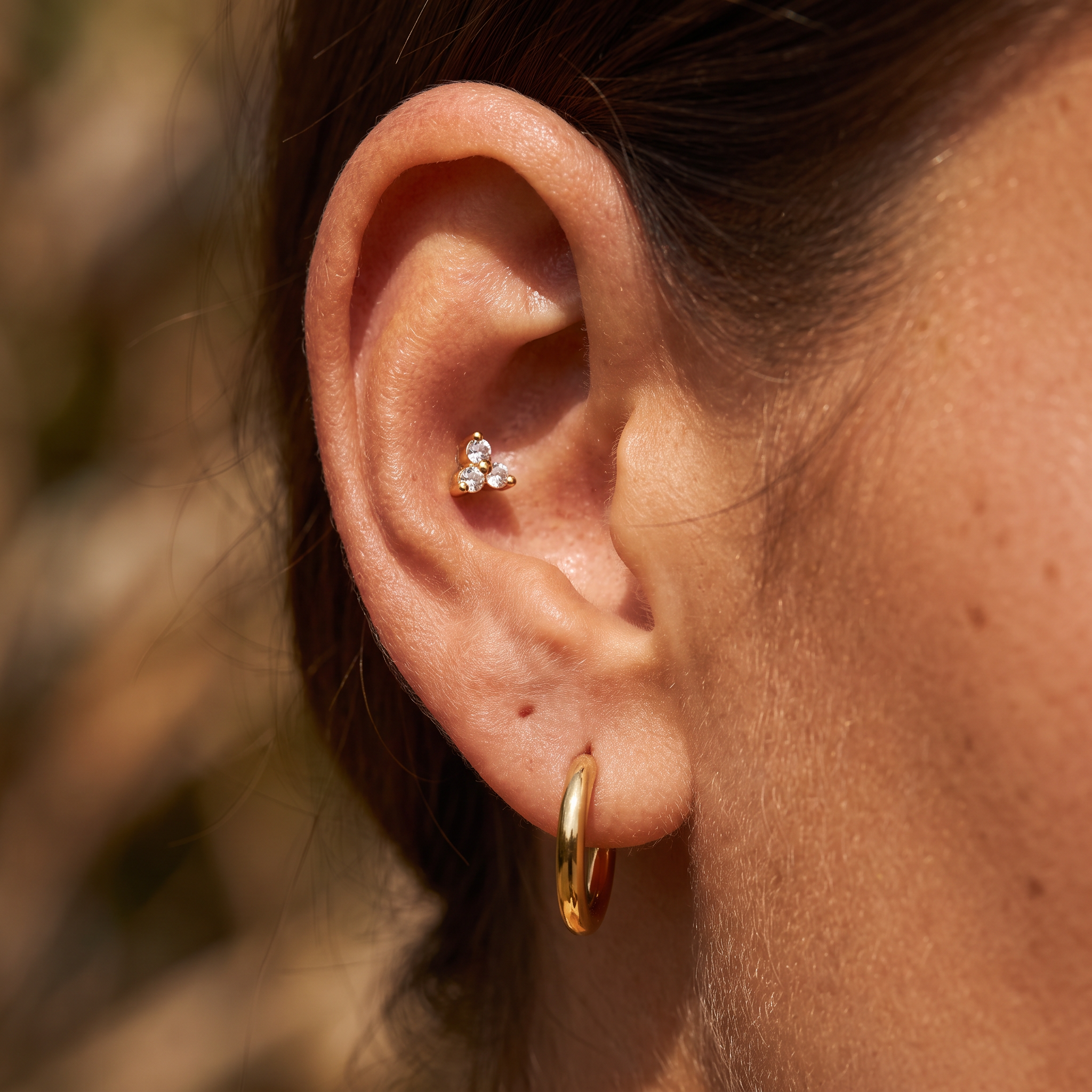 Close-up of a person's ear showcasing a small gold ear cuff with three clear gemstones, paired with a gold hoop earring.