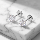A pair of round-cut diamond stud earrings in a silver-toned setting, displayed on a white marble block.
