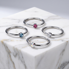Four silver-toned ring bands arranged on a white marble pedestal, each set with a round gemstone in a different color: pink, light blue, clear, and iridescent.
