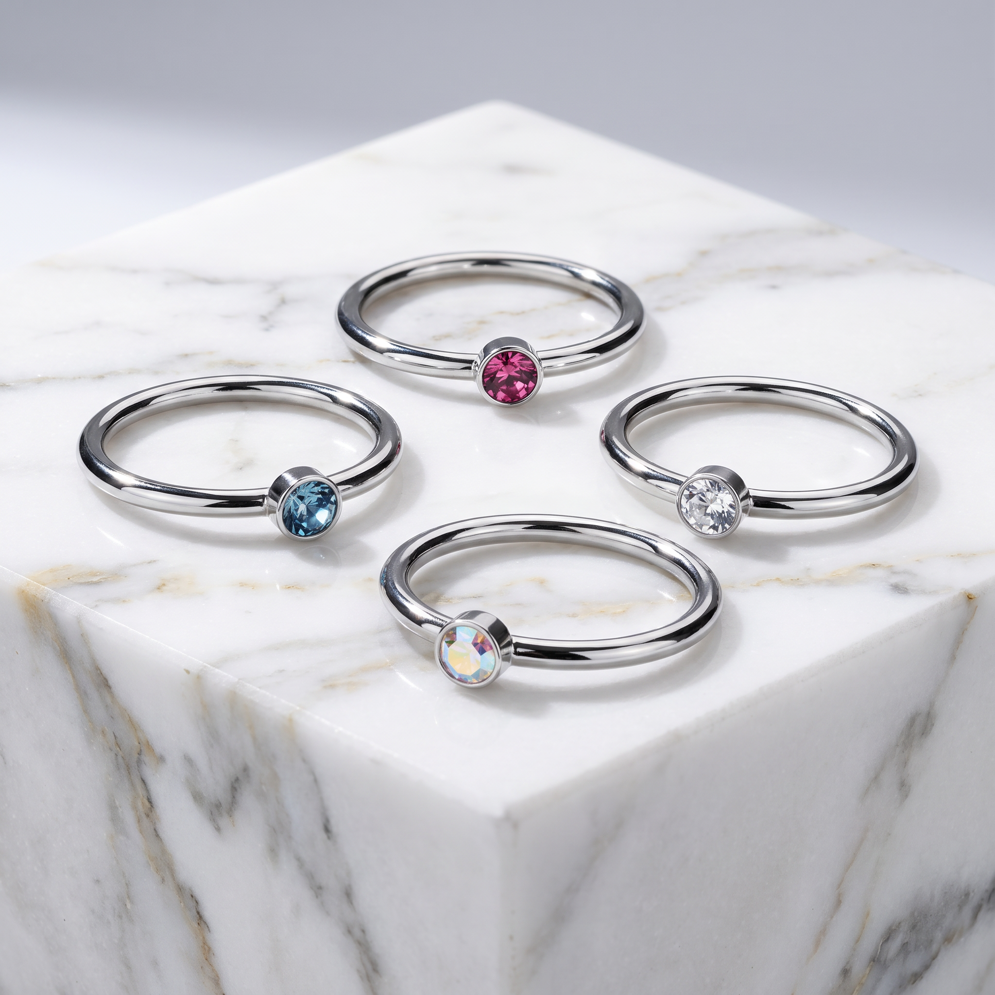 Four silver-toned ring bands arranged on a white marble pedestal, each set with a round gemstone in a different color: pink, light blue, clear, and iridescent.