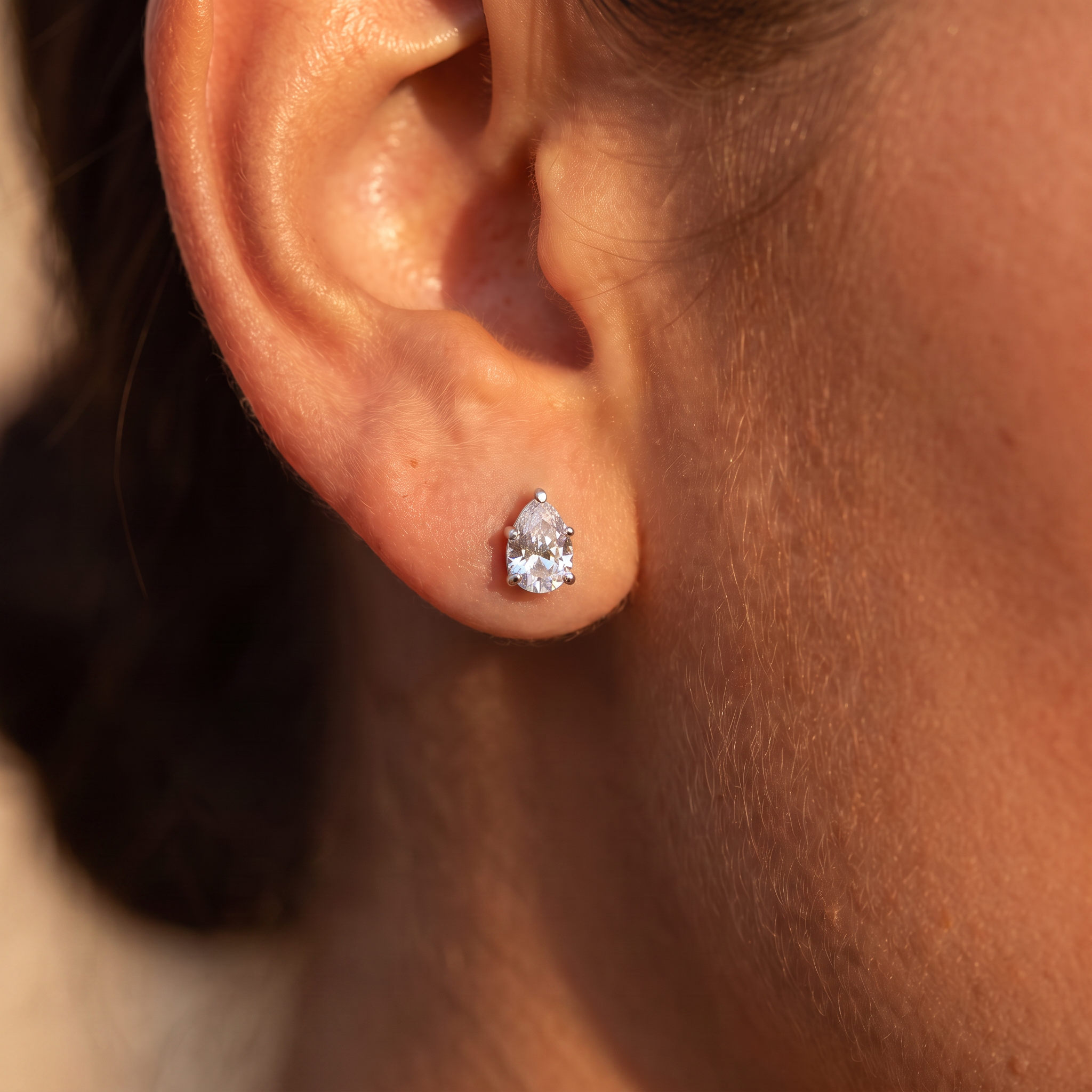 A close-up of a person's ear wearing a small pear-cut clear gemstone stud earring in a silver-toned setting.