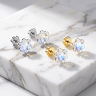 Four flower-shaped stud earrings with iridescent clear gemstones and gold or silver posts, arranged on a white marble slab.