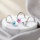 Three silver-toned U-shaped hoop earrings with heart-shaped crystal studs in light blue, pink, and clear, displayed on a white marble pedestal with soft gray fabric in the background.