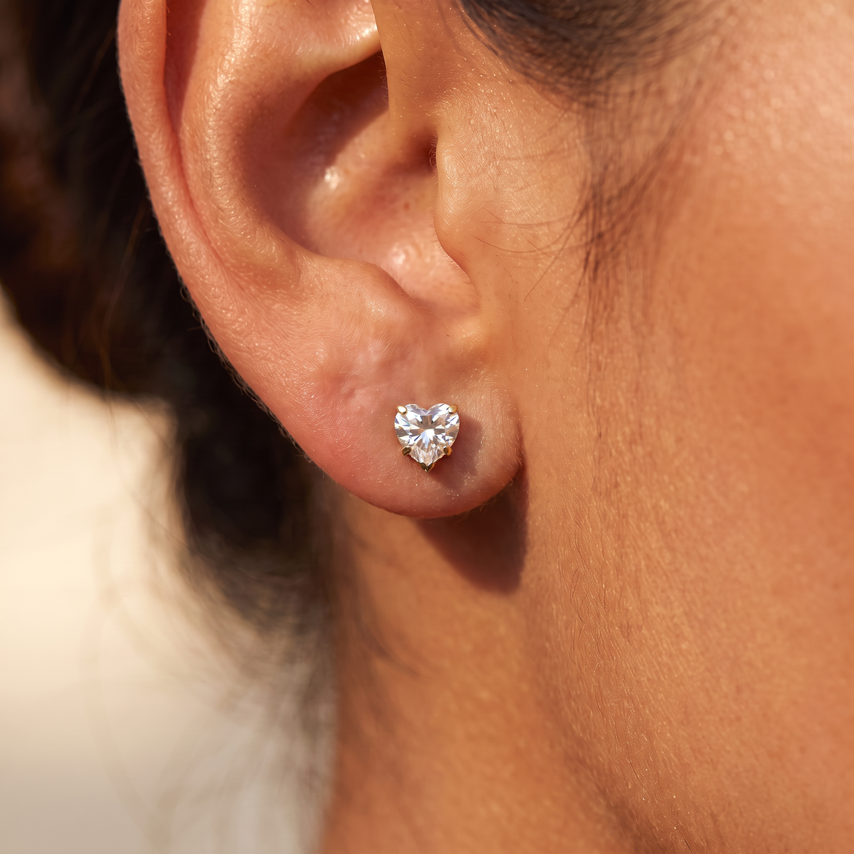 A close-up of a person's ear wearing a small heart-shaped stud earring made of sparkling clear gemstones in a silver-toned setting.
