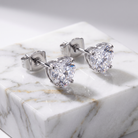 A pair of heart-shaped stud earrings with clear faceted gemstones in a silver-toned setting, displayed on a white marble block against a soft gray background.