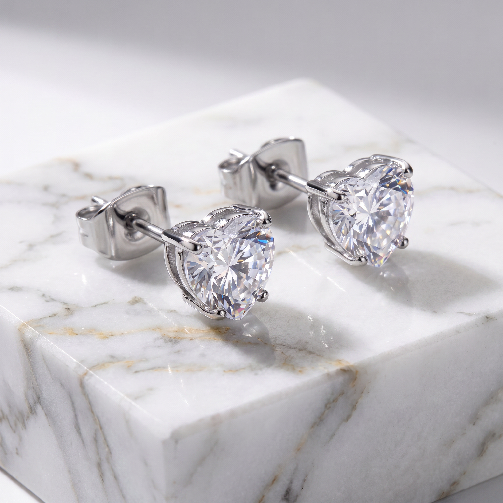 A pair of heart-shaped stud earrings with clear faceted gemstones in a silver-toned setting, displayed on a white marble block against a soft gray background.