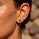 A close-up of a person's ear wearing a small, polished gold heart stud earring, set against a softly blurred natural background.