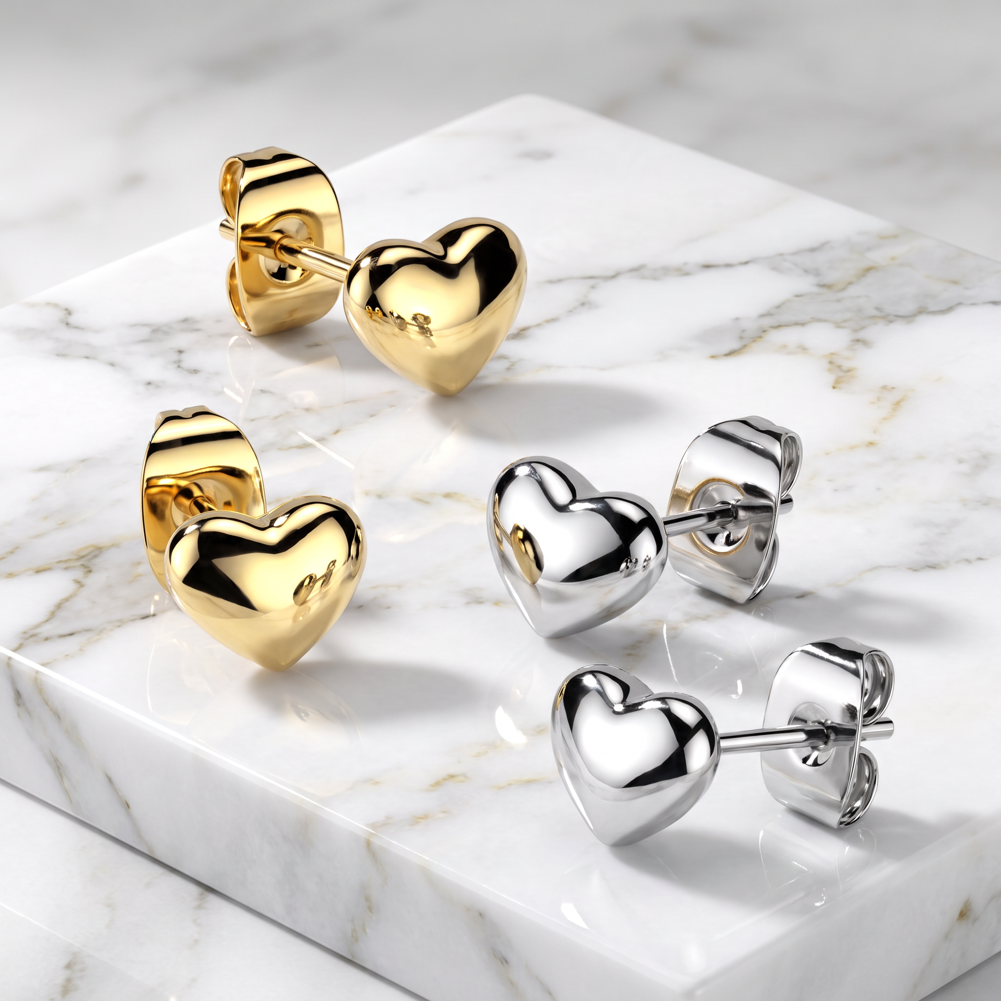 Four heart-shaped stud earrings displayed on a white marble slab, showing both gold and silver finishes with their post and backing details.