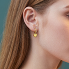 A close-up side profile of a woman wearing a small gold hoop earring with a dangling polished gold heart charm, set against a muted teal background.