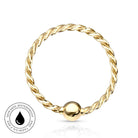 A gold-plated twisted rope ring with a smooth, polished spherical bead at the bottom, displayed on a clean white background with a subtle reflection. A circular badge in the bottom-left corner highlights 'Skin Safe Waterproof' features.