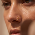 A close-up of a person's face wearing a small twisted gold nose hoop with a bead detail.