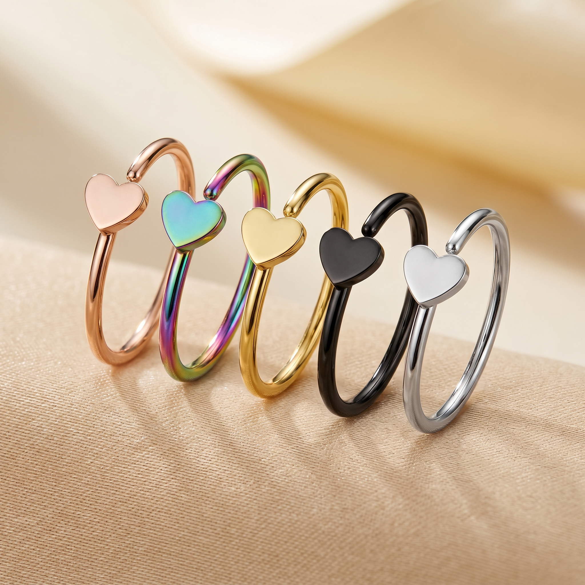 Five slim open bangle rings with heart charms in rose gold, iridescent green, yellow gold, black, and silver, arranged on a beige fabric surface.