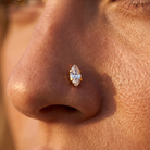 A close-up of a person's nose wearing a small marquise-cut clear gemstone nose stud in a gold setting.