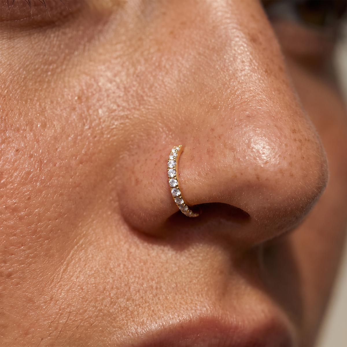 A close-up of a person's nose wearing a delicate gold crescent nose ring adorned with small clear stones.