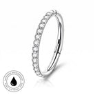 A silver-toned curved ring with a row of clear stones along the front, shown on a white background with a small circular skin-safe waterproof badge in the lower left.