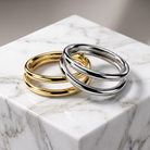 Two polished metal bangle bracelets, one gold-toned and one silver-toned, displayed on a white marble cube against a soft gray background.