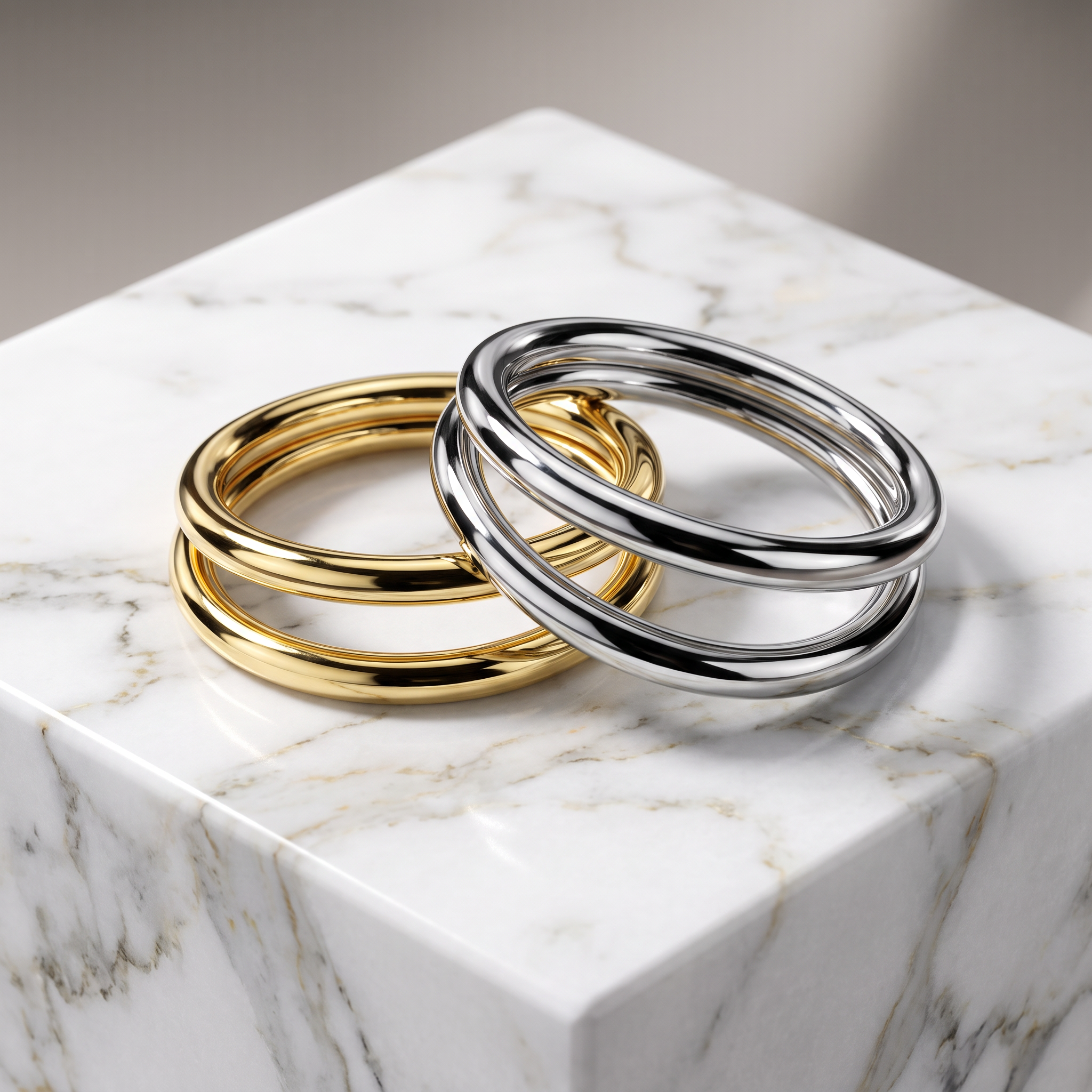 Two polished metal bangle bracelets, one gold-toned and one silver-toned, displayed on a white marble cube against a soft gray background.