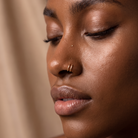 Close-up of a woman with closed eyes wearing a small gold nose ring, against a soft beige background.