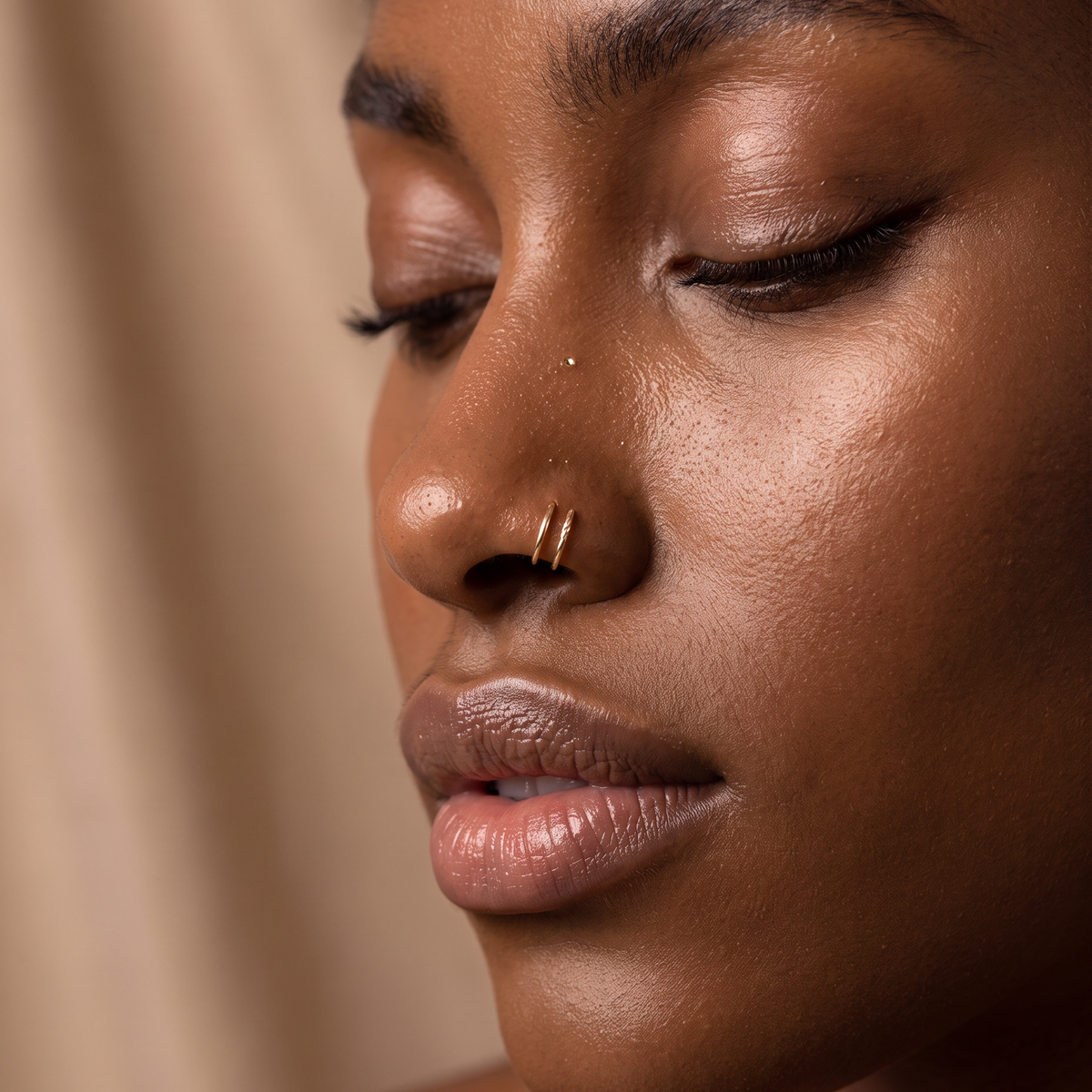 Close-up of a woman with closed eyes wearing a small gold nose ring, against a soft beige background.