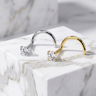 Two curved hoop-style earrings displayed on a white marble block. One earring is silver-toned with a clear round gemstone, and the other is gold-toned with a matching clear round gemstone.