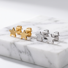 Two pairs of star-shaped stud earrings displayed on a white marble slab. One pair is gold-toned and the other is silver-toned, photographed against a clean white background.