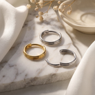 Three minimalist double-band hoop earrings in gold and silver, styled on a white marble slab with soft white fabric and dried flowers.