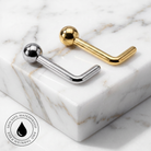 Two curved barbell-style body jewelry pieces with spherical ends, one polished silver and one polished gold, displayed on a white marble block. A circular badge in the lower-left corner reads 'SKIN SAFE WATERPROOF' with a droplet icon.